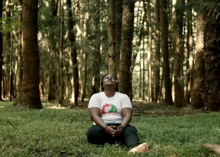 Nzambi Matee sitting on grass in a forest between tress.